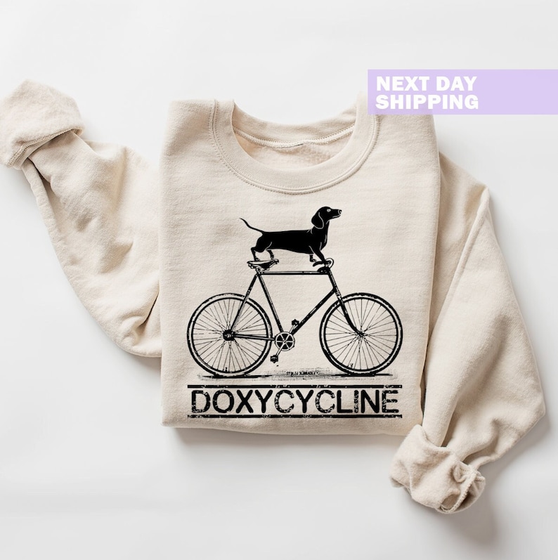 May include: A beige sweatshirt with a black dachshund riding a bicycle and the text "DOXYCYCLINE" printed on the front.