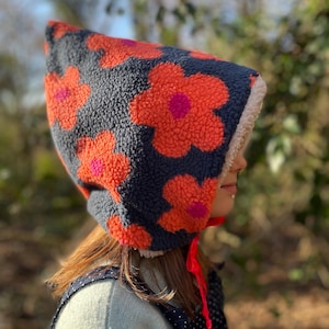 Puede incluir: Un gorro con capucha de fondo azul marino y grandes flores naranjas con centros rosas. El gorro está hecho de un material suave y texturizado y tiene un forro color crema. Cintas rojas brillantes cuelgan del gorro.