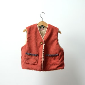 May include: A red corduroy waistcoat with a faux fur lining and a button fastening. The waistcoat has two pockets and a decorative trim around the bottom.