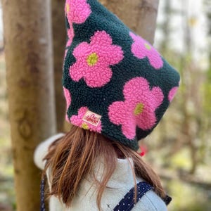 May include: A dark green fleece hat featuring large pink flower patterns with yellow centres. The hat has a pointed shape and a small label that reads "Dee Agnew". This is a clothing accessory.