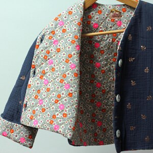 May include: Navy blue quilted jacket with a floral print lining. The jacket features a button-up front and is adorned with gold leaf accents. The interior lining showcases a pattern of pink and orange flowers on a gray background.