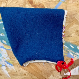 May include: A blue bonnet with a floral patterned lining. The bonnet is made of a soft, textured material and has a tag with a red ribbon that says "Merry Christmas!"