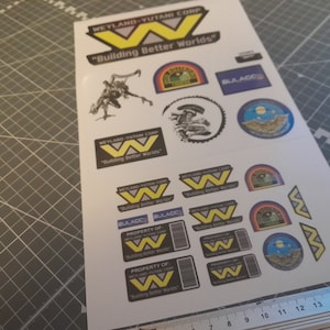 Puede incluir: Hoja de pegatinas con varios diseños de ciencia ficción. Las pegatinas presentan el logotipo de Weyland-Yutani Corp, una "W" amarilla sobre fondo negro, y el lema "Building Better Worlds". Incluye otros gráficos.