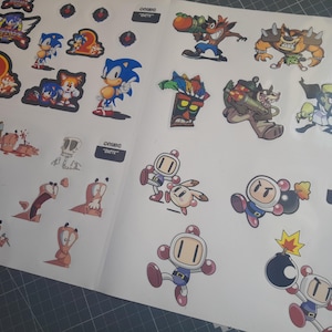 Könnte beinhalten: Ein Bogen mit Aufklebern von Comicfiguren, darunter Sonic the Hedgehog, Crash Bandicoot und Bomberman. Die Aufkleber befinden sich auf weißem Hintergrund. Die Wörter "COMIC" und "QUOTE" sind auf kleinen schwarzen Rechtecken aufgedruckt.