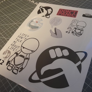 Op de afbeelding: Een vel stickers met zwart-witte illustraties en tekst. Ontwerpen omvatten een robot, een planeet met een hand, een dolfijn en de woorden "DON'T PANIC" in het rood. Het nummer "42" is ook aanwezig.