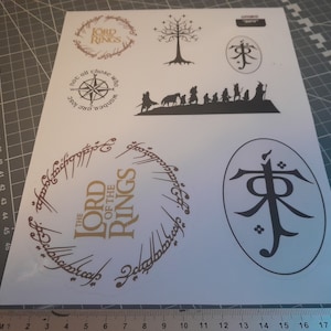 Könnte beinhalten: Ein Bogen mit Aufklebern mit Designs aus "Der Herr der Ringe". Die Aufkleber enthalten den Text "The Lord of the Rings" in einem kreisförmigen Design, einen Kompass und Silhouetten von Charakteren und Symbolen. Die Aufkleber sind in Gold und Schwarz.