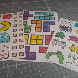 May include: Three sheets of stickers with pixelated designs of classic video game characters. Includes Pac-Man, Tetris blocks, and Space Invaders. The stickers are printed on white paper with black outlines and bright colours.