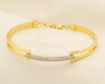 Puede incluir: Una pulsera de tono dorado con una barra central curva incrustada con pequeñas piedras transparentes. La pulsera tiene un diseño de doble banda que se conecta a un cierre de eslabones. El texto STAR LIKES GOLD es visible.
