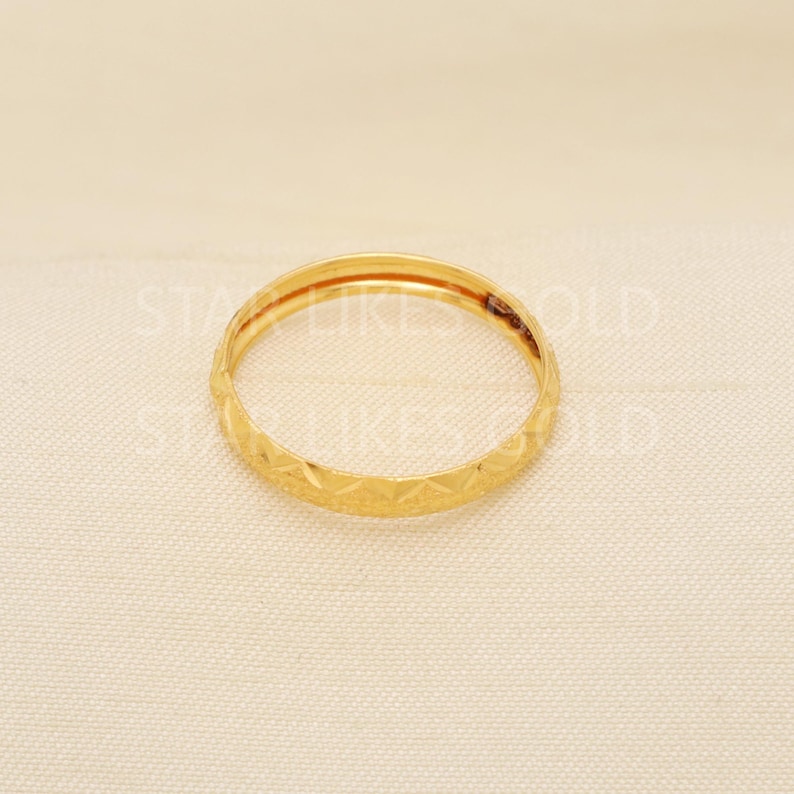 May include: A gold ring with a geometric pattern. The ring is a thin band with a repeating diamond-like design around the exterior. The ring is set against a neutral background.