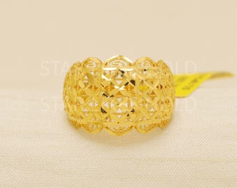 May include: A gold-colored ring with an intricate openwork design. The ring features a repeating pattern of geometric shapes and floral elements. The ring is displayed on a cream-colored surface.