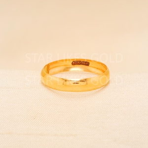 Puede incluir: Una alianza de boda dorada con un perfil redondeado. El anillo tiene una superficie lisa y pulida y una pequeña inscripción. El anillo se muestra sobre una superficie de color neutro. Joyería.