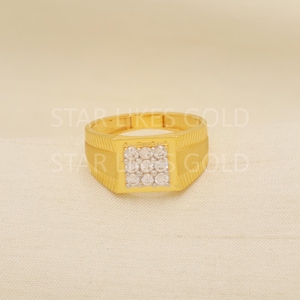 May include: A gold ring featuring a square face adorned with small, clear gemstones. The ring has a textured band and a rectangular face. The ring is presented on a cream-coloured surface.