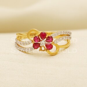 Könnte beinhalten: Ein goldener Ring mit einem roten Blumendesign. Die Blume hat fünf Blütenblätter und einen Mittel-Stein. Der Ring ist mit kleinen weißen Steinen besetzt.