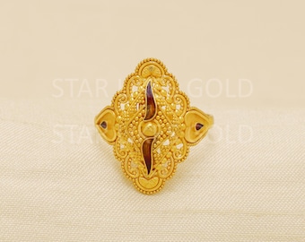 May include: Ornate gold ring with an oval centerpiece. The ring features intricate filigree detailing, a central vertical design with a dark red accent, and small dark red accents on the sides. The words "STAR GOLD" are partially visible.