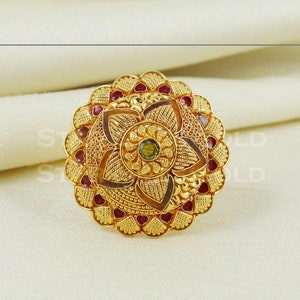 Op de afbeelding: Een gouden ring met een bloemmotief en rode en groene edelstenen. De ring is ingewikkeld en gedetailleerd, met een verhoogd middenstuk en een getextureerde buitenrand.