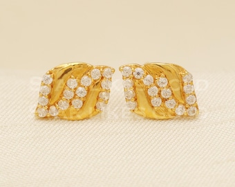 May include: A pair of gold-coloured stud earrings. Each earring has a leaf-like shape with a textured surface and is adorned with multiple small, clear gemstones. The earrings are displayed on a cream-coloured surface.