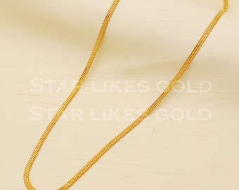May include: A gold chain necklace with a delicate, flat, and slightly textured design. The chain is laid out on a cream-colored surface.