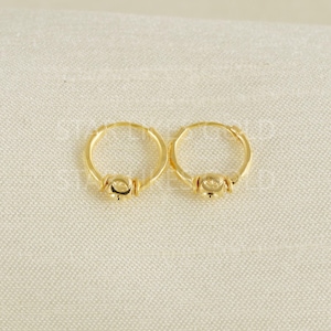 Puede incluir: Un par de pendientes de aro pequeños en color dorado, cada uno con una banda circular lisa y pulida con un detalle de cuenta central en la parte inferior. Los pendientes tienen un acabado metálico brillante y un diseño de aro continuo.