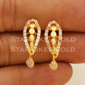 May include: A pair of gold-colored earrings. Each earring features a central design with three stacked teardrop shapes, surrounded by small, clear stones. A teardrop-shaped charm with clear stones hangs from the bottom. The text "STAR" and "GOLD" is visible.