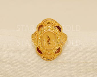 May include: A gold-colored ring with an oval centerpiece and ornate detailing. The ring features a central design with a small, dark accent and is surrounded by a decorative border. The ring has a floral-like design with red accents.