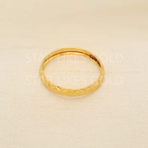 May include: A gold ring with a geometric pattern. The ring is a thin band with a repeating diamond-like design around the exterior. The ring is set against a neutral background.
