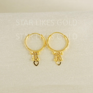 Könnte beinhalten: Ein Paar goldene Creolen mit hängenden Kettenelementen und einem kleinen Herzanhänger an der Basis jedes Rings. Der Text STAR LIKES GOLD ist im Hintergrund sichtbar. Die Ohrringe haben eine polierte Oberfläche und ein rundes Design.