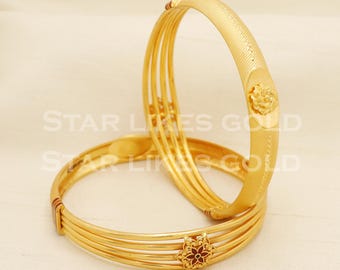 Könnte beinhalten: Zwei goldene Armreifen im mehrsträngigen Design mit polierter Oberfläche. Ein Armreif zeigt eine texturierte Oberfläche mit einem zentralen Blumenmotiv, während der andere einen kleinen, detaillierten Blumenakzent mit dunkelroter Emaille aufweist.