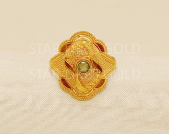 May include: A gold-colored ring with an ornate design. The ring features a central green gemstone and red accents. The ring has a detailed, textured surface and is set against a neutral background.