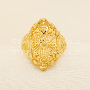 Könnte beinhalten: Ein goldfarbener Ring mit einem verzierten, strukturierten Design. Der Ring hat ein zentrales rautenförmiges Element mit komplizierten Mustern und herzförmigen Akzenten. Der Ring ist vor einem neutralen Hintergrund platziert.