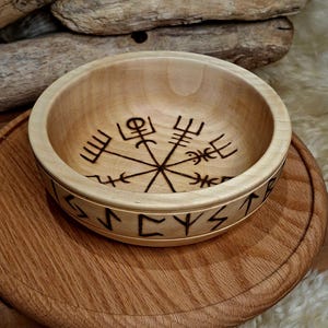 May include: A light-coloured wooden bowl featuring a dark, burned-in Vegvisir symbol at the centre of the interior base. The exterior side of the bowl is decorated with a continuous band of dark, burned-in runic characters encircling the rim.