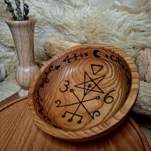 May include: A round wooden bowl with a natural light brown grain, featuring a dark, burned-in design on the interior. The engraving includes a central five-pointed star, geometric lines, the numbers 3, 6, 2, 4, and a crescent moon symbol.