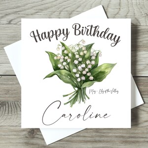 May include: A white card with the text "Happy Birthday" in black cursive font. A bouquet of white lily of the valley flowers with green leaves is in the center of the card. The text "May - Lily of the Valley" is below the flowers. The name "Caroline" is written in cursive at the bottom of the card.