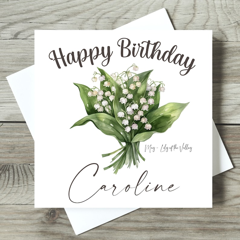 May include: A white card with the text "Happy Birthday" in black cursive font. A bouquet of white lily of the valley flowers with green leaves is in the center of the card. The text "May - Lily of the Valley" is below the flowers. The name "Caroline" is written in cursive at the bottom of the card.