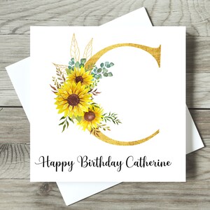 May include: A white birthday card featuring a gold letter "C" decorated with sunflowers and foliage. The card says "Happy Birthday Catherine" in black script. The card is on a white envelope.