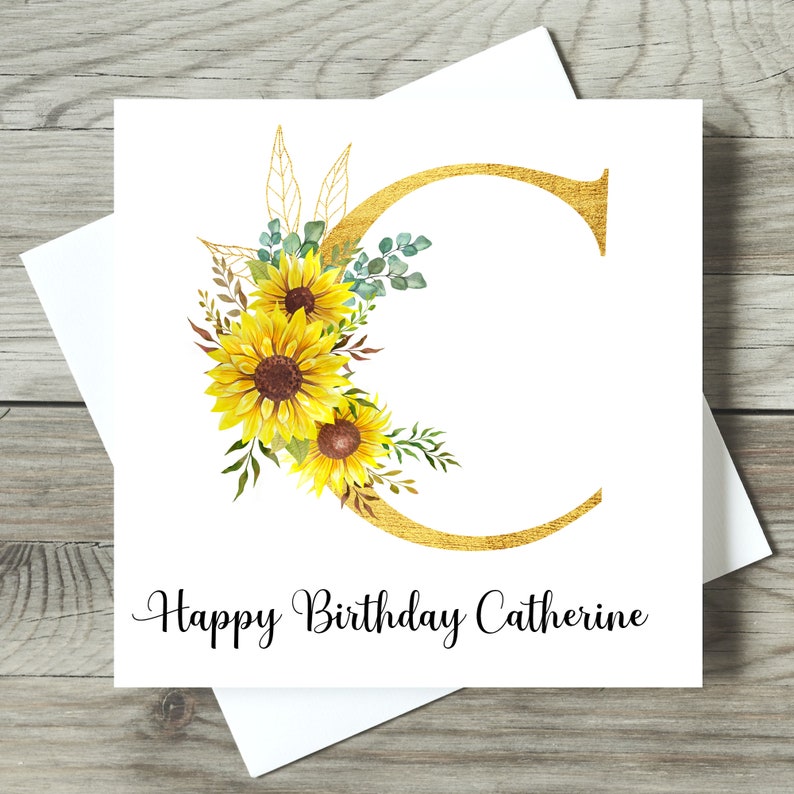 May include: A white birthday card featuring a gold letter "C" decorated with sunflowers and foliage. The card says "Happy Birthday Catherine" in black script. The card is on a white envelope.