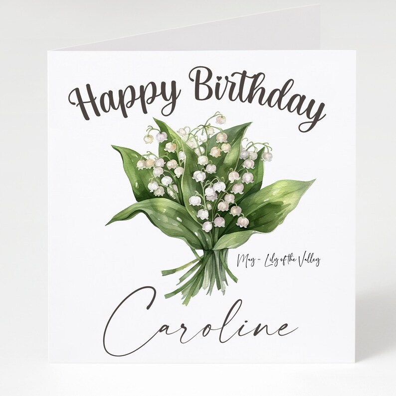 May include: A white card with the text "Happy Birthday" in brown cursive lettering. A bouquet of white lily of the valley flowers with green leaves is in the center of the card. The text "May - Lily of the Valley" is below the flowers. The name "Caroline" is written in brown cursive lettering at the bottom of the card.
