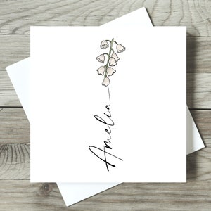 May include: A white card with a simple line drawing of a lily of the valley flower and the name Amelia written in cursive.