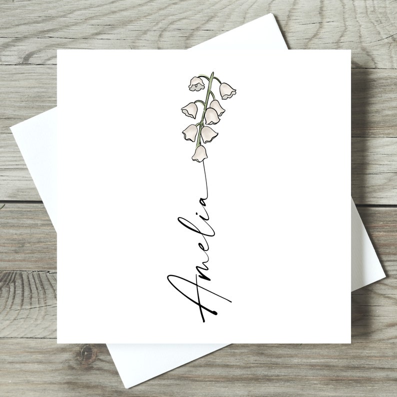 May include: A white card with a simple line drawing of a lily of the valley flower and the name Amelia written in cursive.