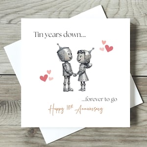 May include: A white greeting card with a drawing of two robot figures holding hands, surrounded by pink hearts. The card reads "Tin years down...forever to go" and "Happy 10th Anniversary" in a handwritten style font.