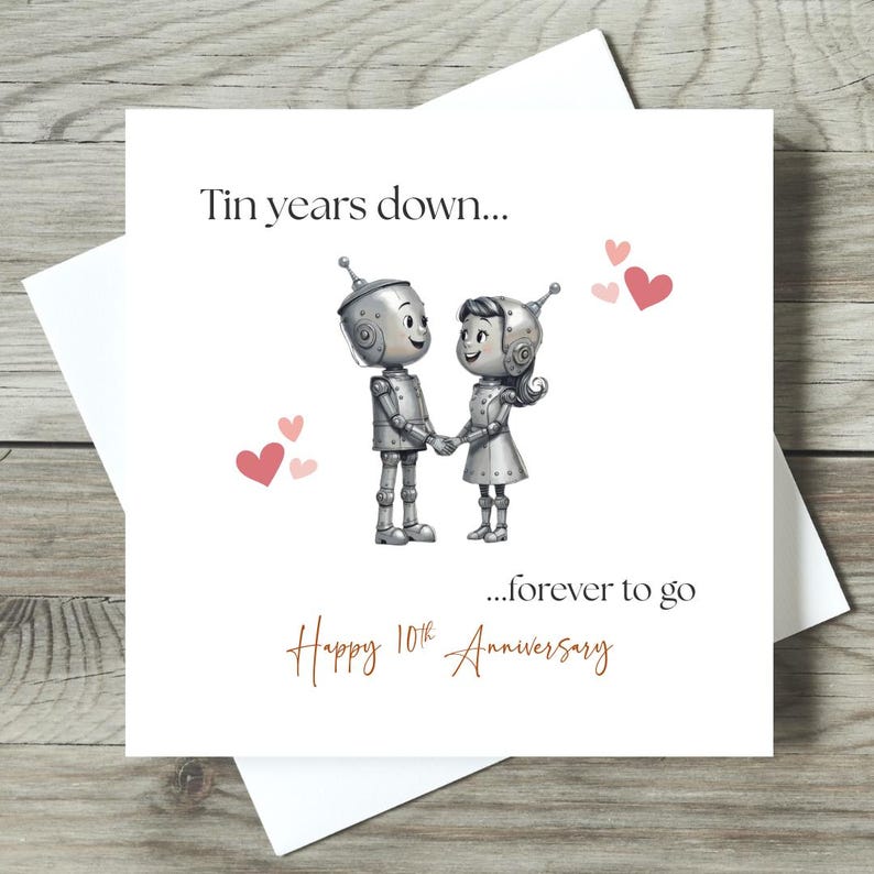 May include: A white greeting card with a drawing of two robot figures holding hands, surrounded by pink hearts. The card reads "Tin years down...forever to go" and "Happy 10th Anniversary" in a handwritten style font.