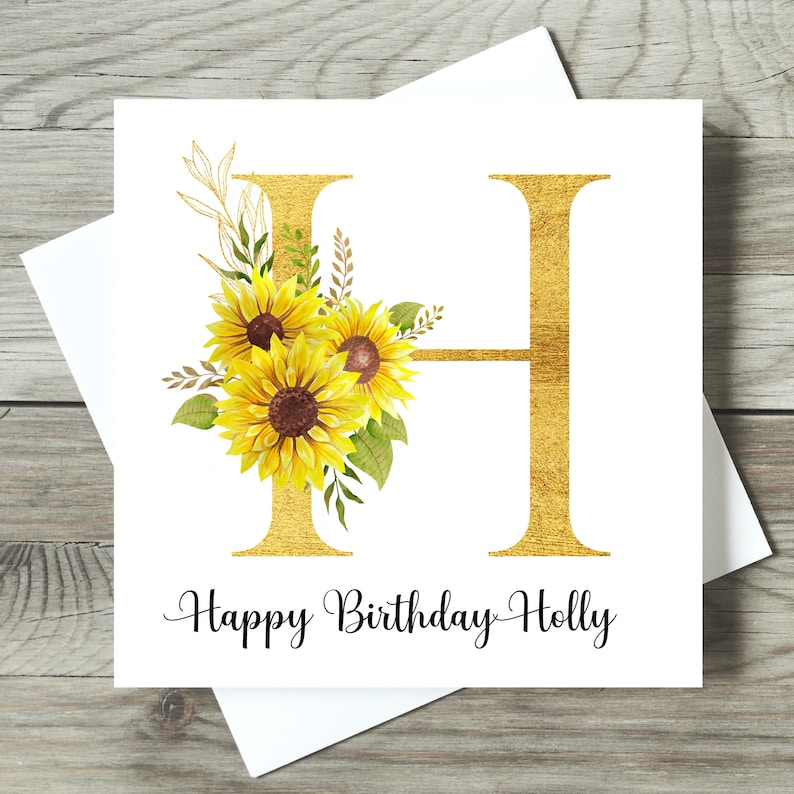 May include: A white birthday card featuring a gold glitter letter 'H' and the words 'Happy Birthday Holly'. The letter is adorned with yellow sunflowers and green foliage. The card is presented with a white envelope.