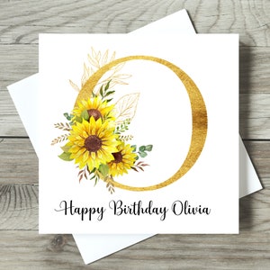 May include: A white birthday card featuring a gold-coloured letter "O" and a floral arrangement of yellow sunflowers. The card says "Happy Birthday Olivia" in black script. The card is on a wooden surface.