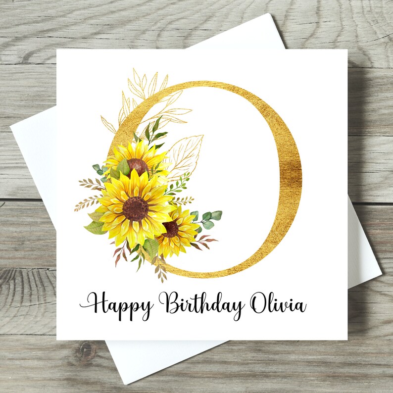 May include: A white birthday card featuring a gold-coloured letter "O" and a floral arrangement of yellow sunflowers. The card says "Happy Birthday Olivia" in black script. The card is on a wooden surface.
