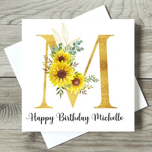 May include: A square birthday card featuring a gold letter "M" adorned with sunflowers and foliage. The words "Happy Birthday Michelle" are written in black script below. The card is presented with a white envelope.