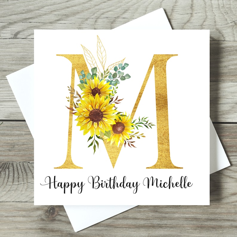 May include: A square birthday card featuring a gold letter "M" adorned with sunflowers and foliage. The words "Happy Birthday Michelle" are written in black script below. The card is presented with a white envelope.