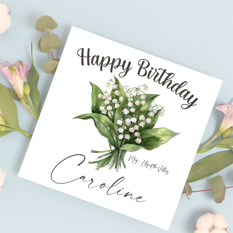 May include: A white card with the text "Happy Birthday Caroline" and a bouquet of white lily of the valley flowers. The text "May - Lily of the Valley" is written below the flowers.