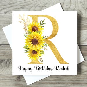 May include: A white birthday card featuring a gold letter "R" decorated with yellow sunflowers and green foliage. The words "Happy Birthday Rachel" are written in black script. The card is set against a white backdrop.