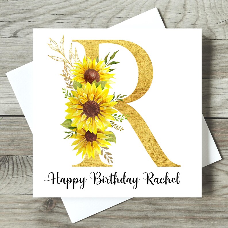 May include: A white birthday card featuring a gold letter "R" decorated with yellow sunflowers and green foliage. The words "Happy Birthday Rachel" are written in black script. The card is set against a white backdrop.