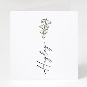 May include: A white card with a simple line drawing of a lily of the valley flower and the name "Hayley" written in cursive.