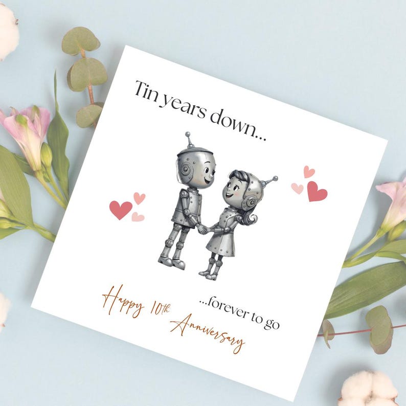 May include: A white anniversary card with the text "Tin years down..." and "...forever to go". The card features a cartoon illustration of two robots holding hands, with pink hearts scattered around them. The words "Happy 10th Anniversary" are written in gold script.